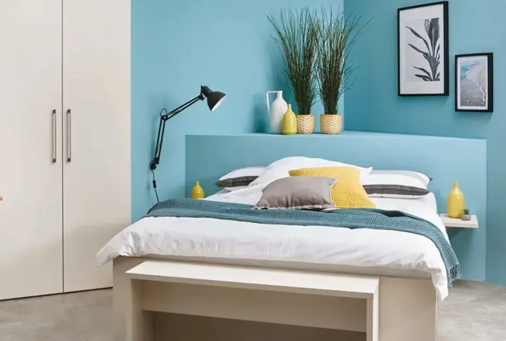 Weisses Bett mit Kissen in Gelb und Grau vor blauer Wand, links weisser Kleiderschrank, auf dem Kopfteil Vasen mit Gräsern, rechts zwei gerahmte Bilder.